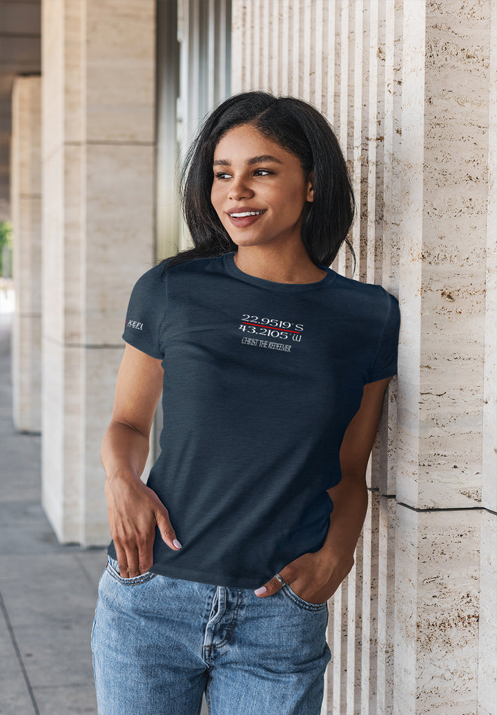A confident woman in a blue t-shirt leans against a wall with her hands on her hips, exuding composure and casual grace.