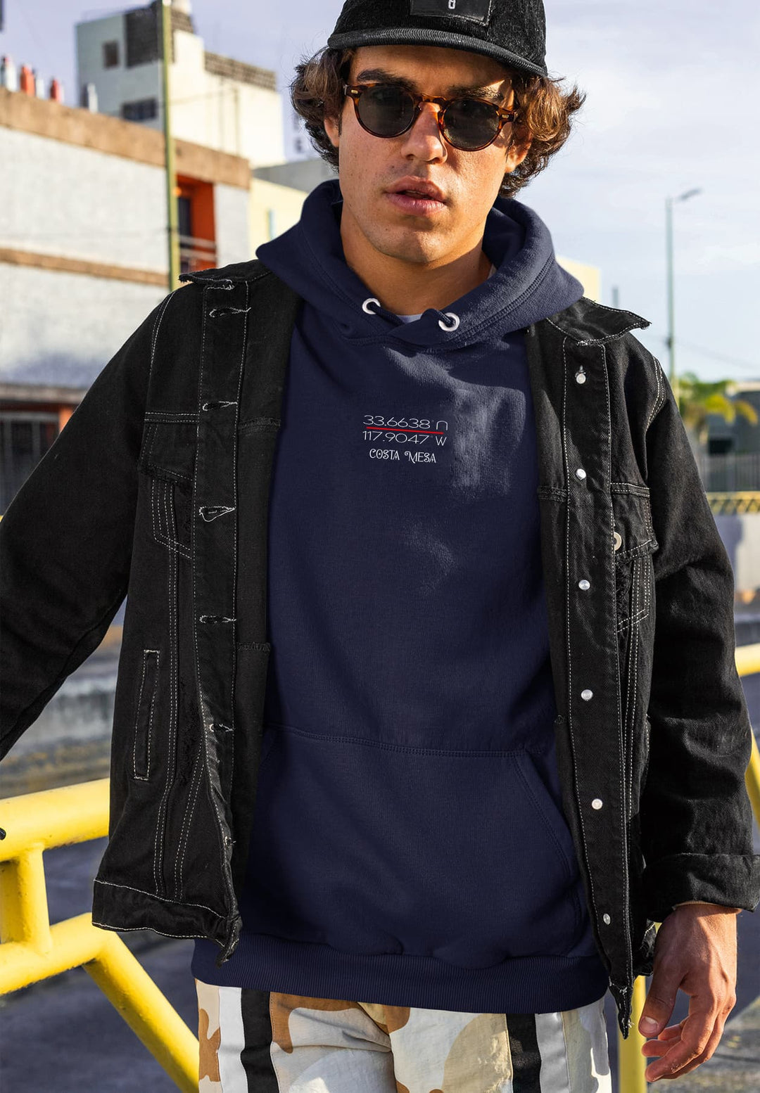 A guy wearing a black jacket over a navy blue hoodie standing by a street rail.