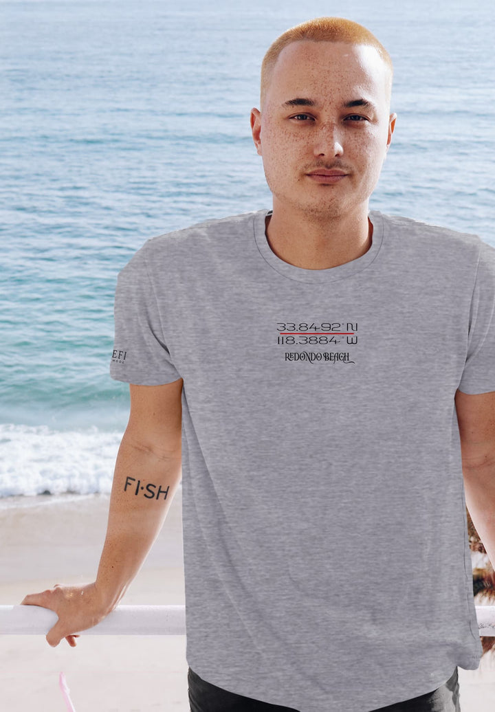 The hipster man stands by the sea, his gray coordinate t-shirt blending with the serene coastal palette.