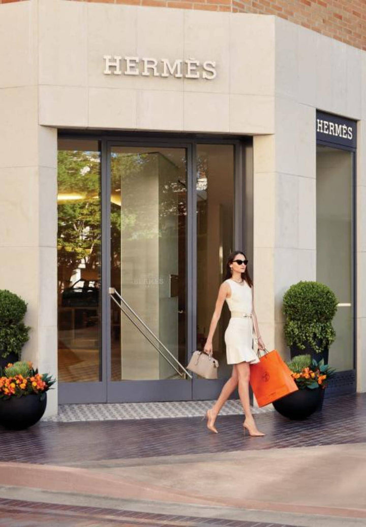 A woman adorned in a fashionable garment departs from the shopping center, clutching numerous packages, exuding the delight of discovering newfound treasures.