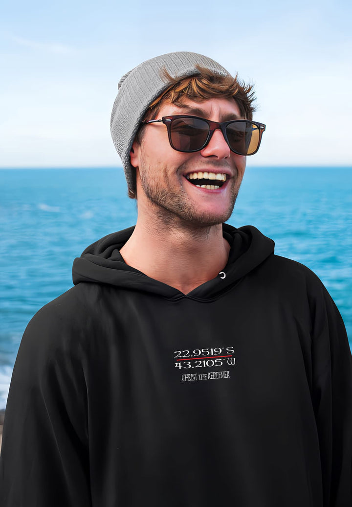 A man dressed in a black hoodie, sunglasses, and a hat while standing by the ocean.