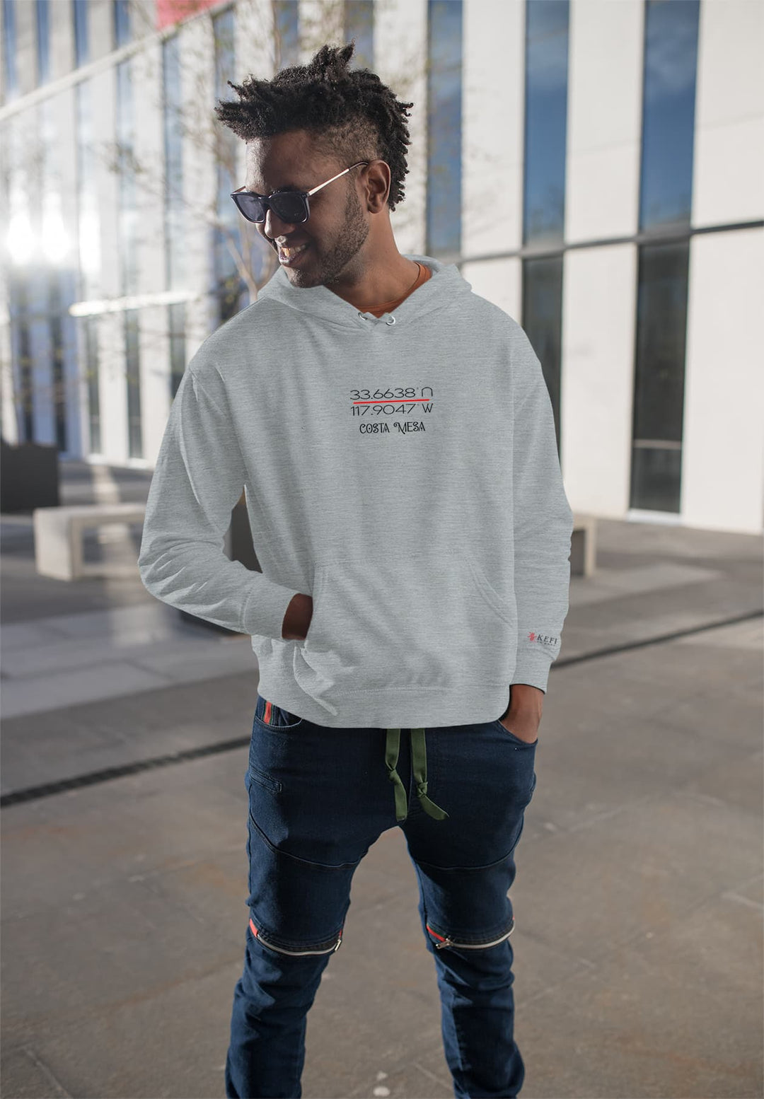 A male with dreadlocks and sunglasses standing on a sidewalk wearing a men's gray hoodie looking off to the side.