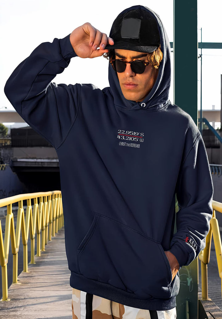 A gentleman walks down the street, donning a hat, sunglasses, and a navy blue hoodie.