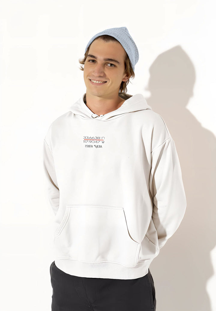 A young man sporting a white hoodie with a Costa Mesa design and standing in the middle of a studio.