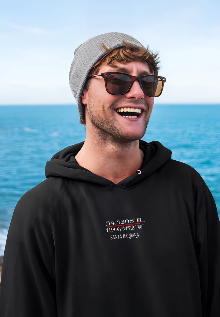 The man wears a Santa Barbara black hoodie by the seaside with elegance and contemporary fashion.