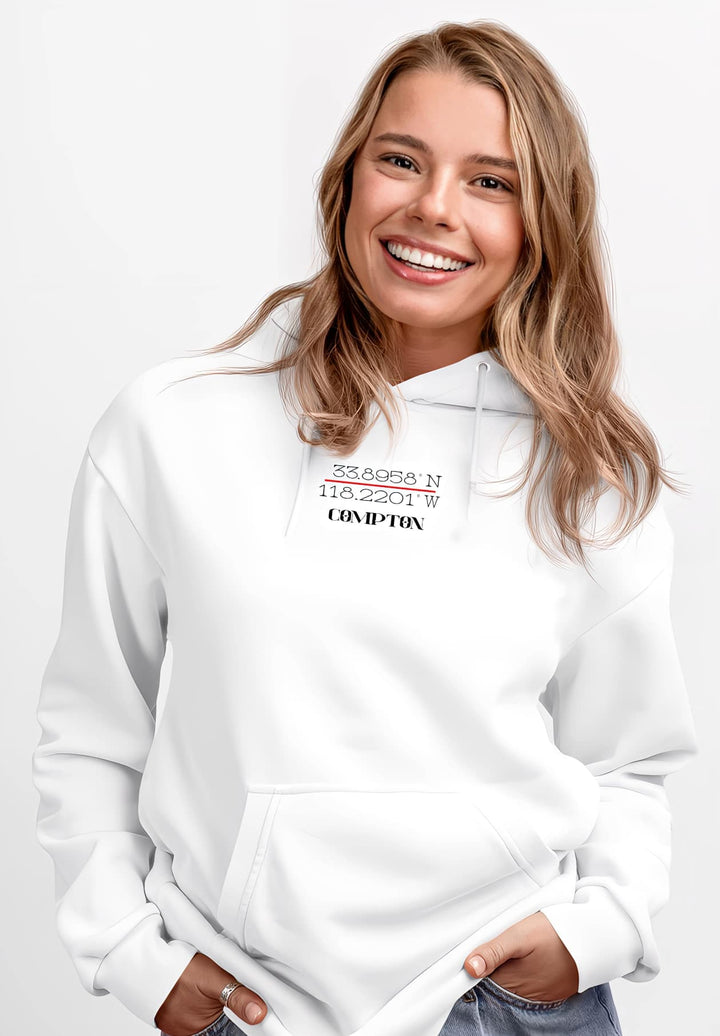 A woman who is displaying a smile is attired in a white hoodie garment that features GPS coordinates of Compton, California.