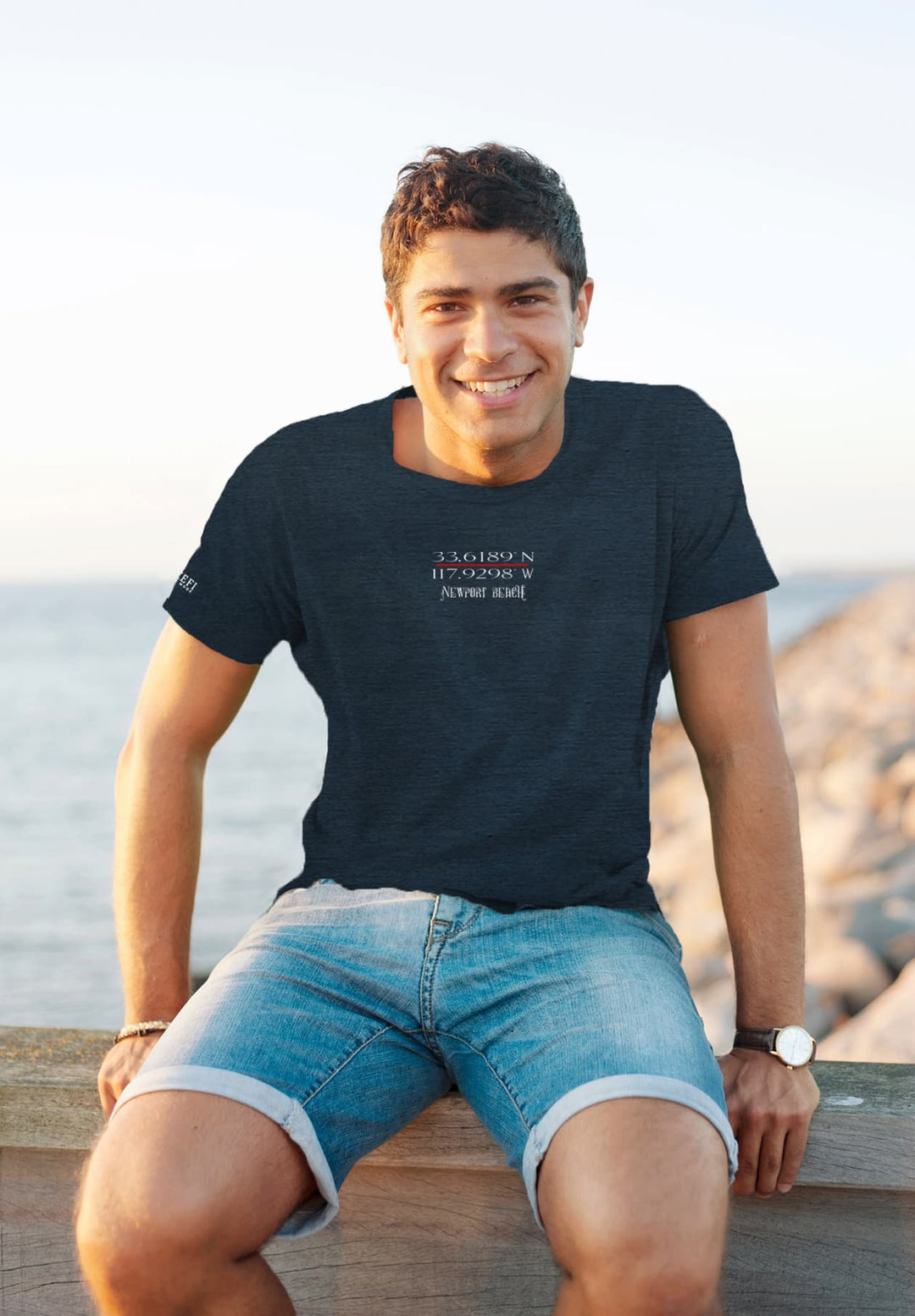 By the ocean's serenity, the smiling man in a blue tee radiates joy, his happiness mirroring the endless horizon.