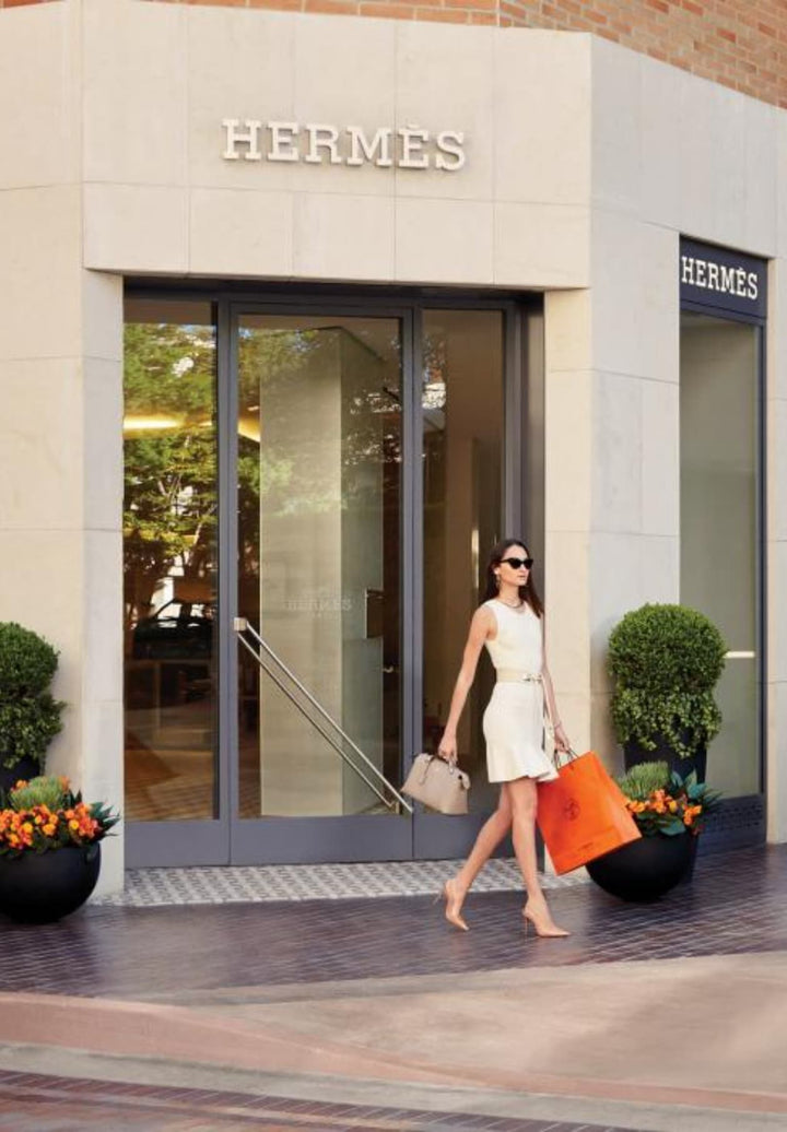 A shopper seen exiting the South Coast Plaza shopping center wearing a white dress and carrying shopping bags in heels.
