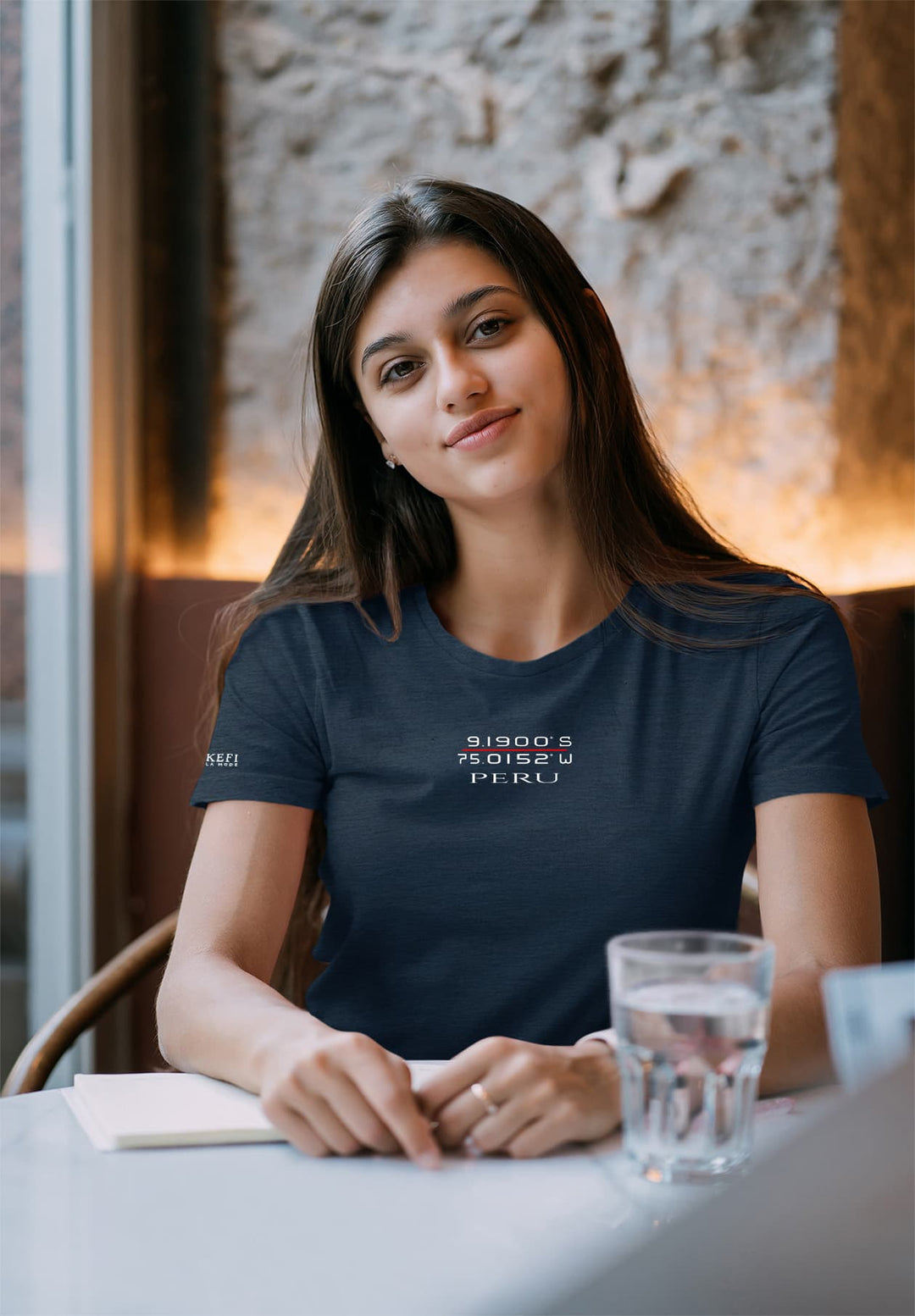 In the soft glow, she sits, a tranquil moment painted in blue t-shirt, a serene tableau of contemplation with a glass of water.
