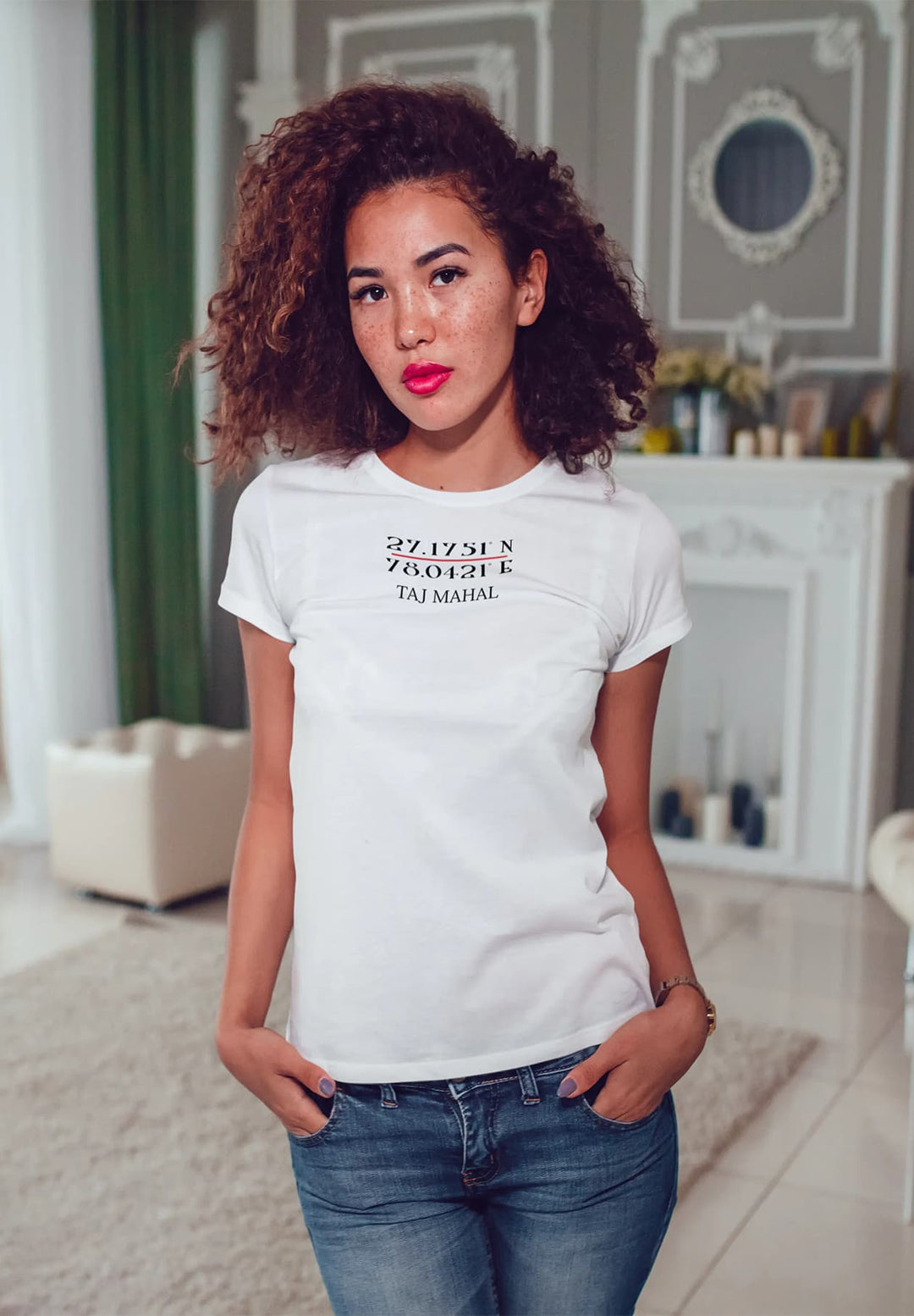Effortlessly cool, she stands in her living room, hands casually in pockets, wearing a white T-shirt, embodying a relaxed and stylish vibe.