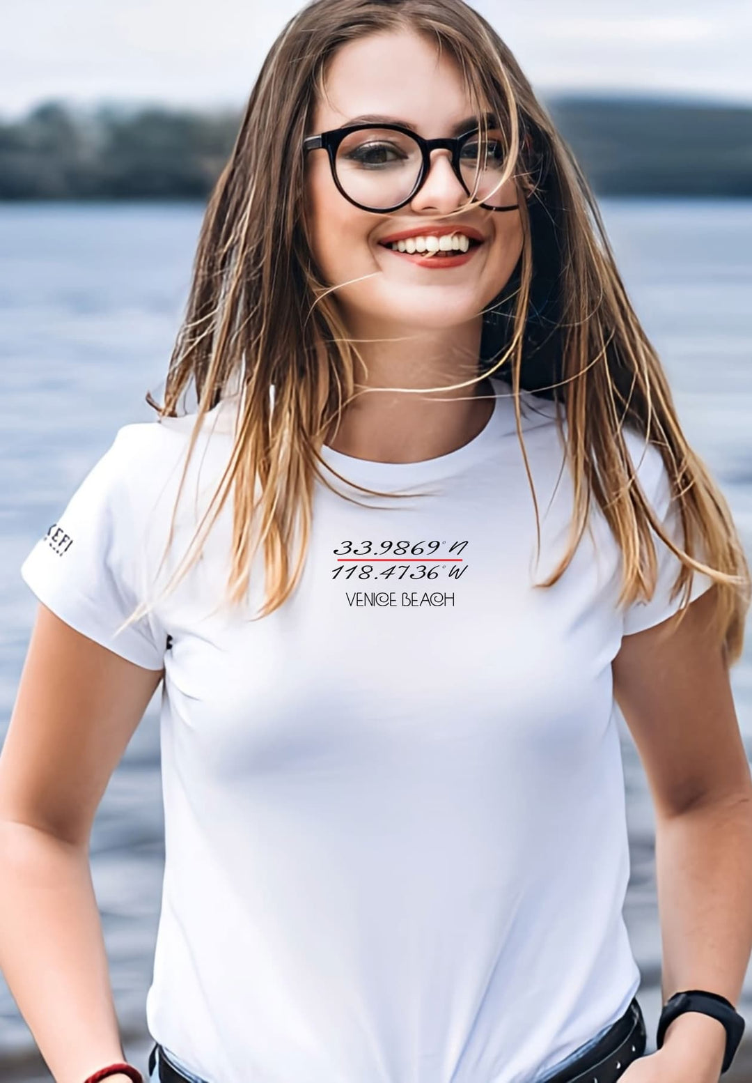 Beneath the seaside sky, she wears glasses and a white tee, expressing a cheerful beach style.